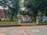 A landscaped outdoor area with large trees and neatly arranged flower beds. A small structure with a red roof sits in the background. There is a decorative design on the paved ground in the foreground, and blue letters spelling out 'ICIT ASD2 23' are displayed prominently.