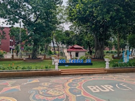 A landscaped outdoor area with large trees and neatly arranged flower beds. A small structure with a red roof sits in the background. There is a decorative design on the paved ground in the foreground, and blue letters spelling out 'ICIT ASD2 23' are displayed prominently.