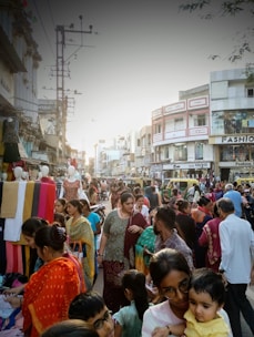 A busy street market filled with people shopping and browsing various stalls. Colorful fabrics and garments are displayed prominently, and the crowd consists of men, women, and children. Surrounding buildings have signs in a local language, contributing to the bustling atmosphere.