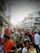 A busy street market filled with people shopping and browsing various stalls. Colorful fabrics and garments are displayed prominently, and the crowd consists of men, women, and children. Surrounding buildings have signs in a local language, contributing to the bustling atmosphere.