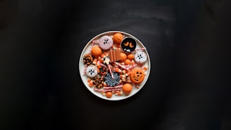 A white plate filled with Halloween-themed treats, including decorated cookies with spooky designs, various candies such as candy corn, candy sticks, and small chocolate pieces. The designs on the cookies include jack-o'-lantern faces, ghost, spider web, and other Halloween motifs. The background is a dark surface that contrasts with the bright colors of the candies.
