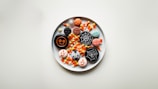 A white plate holds a variety of Halloween-themed treats, including candy corn, macarons decorated with spooky designs such as spiders, webs, and faces, as well as cookies with Halloween patterns.