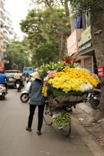 A vendor is pushing a bicycle loaded with a large, colorful arrangement of flowers down a tree-lined street in an urban setting. The flowers include bright yellow, white, and red blooms, and the scene is bustling with various vehicles like scooters and cars nearby. Trees and buildings line the street.