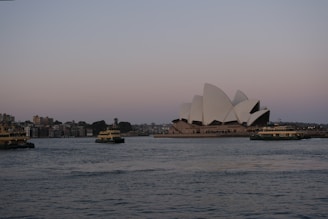 The image depicts a view of the iconic Sydney Opera House sitting on the edge of the harbor during twilight. Several ferries are visible in the water, and a cityscape is seen in the background. The architectural curves of the Opera House contrast against the calm water and the soft hues of the sky.