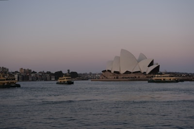 The image depicts a view of the iconic Sydney Opera House sitting on the edge of the harbor during twilight. Several ferries are visible in the water, and a cityscape is seen in the background. The architectural curves of the Opera House contrast against the calm water and the soft hues of the sky.