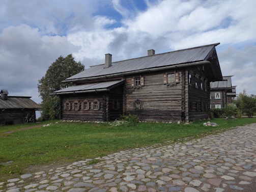 A rustic wooden house with a log cabin design is surrounded by green grass and cobblestone paths. The sky is partly cloudy, casting a soft light on the scene. Trees are visible to the left and right of the house, enhancing the natural and tranquil setting.