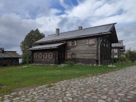 A rustic wooden house with a log cabin design is surrounded by green grass and cobblestone paths. The sky is partly cloudy, casting a soft light on the scene. Trees are visible to the left and right of the house, enhancing the natural and tranquil setting.