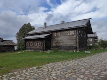 A rustic wooden house with a log cabin design is surrounded by green grass and cobblestone paths. The sky is partly cloudy, casting a soft light on the scene. Trees are visible to the left and right of the house, enhancing the natural and tranquil setting.