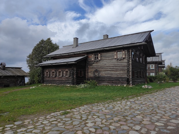 A rustic wooden house with a log cabin design is surrounded by green grass and cobblestone paths. The sky is partly cloudy, casting a soft light on the scene. Trees are visible to the left and right of the house, enhancing the natural and tranquil setting.