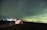 A starry night sky over a serene landscape with a brightly illuminated tent on the ground. The tent is set up near a body of water, reflecting the lights in the background. The sky is filled with numerous stars, creating a sense of vastness and tranquility.