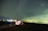 A starry night sky over a serene landscape with a brightly illuminated tent on the ground. The tent is set up near a body of water, reflecting the lights in the background. The sky is filled with numerous stars, creating a sense of vastness and tranquility.