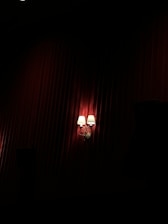 Two wall-mounted lamps with white shades emit a warm glow against a background of deep red, textured drapery. The lighting creates dramatic shadows, enhancing the luxurious ambiance.