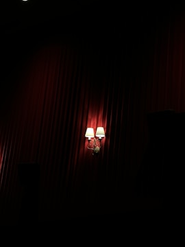 Two wall-mounted lamps with white shades emit a warm glow against a background of deep red, textured drapery. The lighting creates dramatic shadows, enhancing the luxurious ambiance.