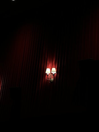 Two wall-mounted lamps with white shades emit a warm glow against a background of deep red, textured drapery. The lighting creates dramatic shadows, enhancing the luxurious ambiance.