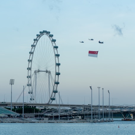 A large Ferris wheel stands prominently near a body of water, surrounded by modern infrastructure. In the sky, four helicopters fly in formation, with one carrying the national flag of Singapore. The scene is set against a clear blue sky at dusk.