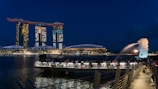 A waterfront scene at night featuring a large, illuminated hotel with three connected towers and a boat-like structure on top. The iconic Merlion statue spouts water into the bay, and a crowd is gathered on a boardwalk overlooking the water.