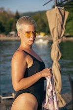 A person with short hair is wearing a black swimsuit and yellow-tinted sunglasses. They are holding a patterned cloth while standing near a body of water. In the background, there are blurred trees and structures, and a curtain-like fabric is draped to the right.