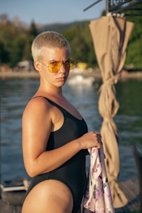 A person with short hair is wearing a black swimsuit and yellow-tinted sunglasses. They are holding a patterned cloth while standing near a body of water. In the background, there are blurred trees and structures, and a curtain-like fabric is draped to the right.
