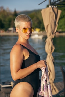 A person with short hair is wearing a black swimsuit and yellow-tinted sunglasses. They are holding a patterned cloth while standing near a body of water. In the background, there are blurred trees and structures, and a curtain-like fabric is draped to the right.