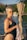 A person with short hair is wearing a black swimsuit and yellow-tinted sunglasses. They are holding a patterned cloth while standing near a body of water. In the background, there are blurred trees and structures, and a curtain-like fabric is draped to the right.