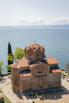 A historic church with red-tiled roofs and intricate brickwork stands on a slope overlooking a vast, tranquil body of water. Surrounding the church are a few tall, slender trees and some green foliage. The background reveals a distant mountain range under a partly cloudy sky.