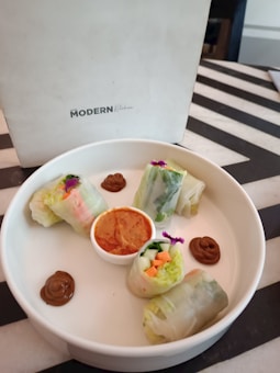 A white plate containing four vegetable spring rolls, each cut in half, arranged around a small dipping sauce bowl. The fillings include assorted vegetables like carrots and lettuce. Swirls of a brown sauce are placed around the rolls. The background features a menu with the words 'Modern Kitchen' on a black and white striped table.