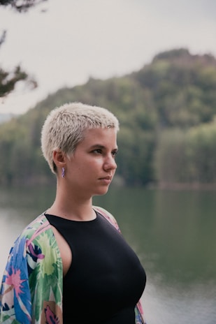 A person with short, light-colored hair gazes thoughtfully into the distance. They are wearing a black top and a colorful, patterned shawl or jacket. The background features a serene body of water and lush, green forested hills.