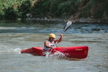 A person is kayaking vigorously in a river, creating splashes as they paddle through the water. The person is wearing a yellow helmet and a sleeveless top, signifying engagement in a water sport. The kayak is red with some writing on it, and the river is surrounded by greenery.