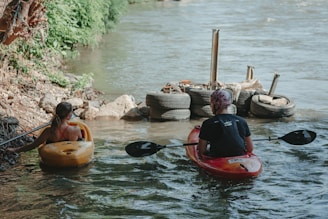 Two individuals are seated in kayaks on a river, preparing to paddle. The person on the left is in a yellow kayak, while the person on the right is in a red kayak. Both are close to the riverbank, which is lined with green foliage and rocks. Ahead of them, tires are stacked by the water's edge.
