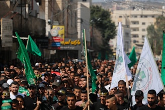 A large crowd of people gathers in a street, holding green and white flags with specific emblems and Arabic script. The scene is densely packed, with individuals wearing dark-colored clothing, some wearing hats or headbands. Buildings with signs in various languages line the street, and the image has a sense of movement and intensity.