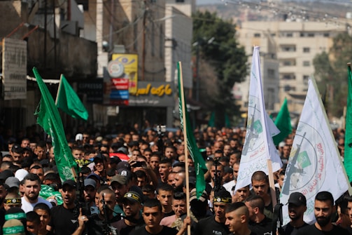 A large crowd of people gathers in a street, holding green and white flags with specific emblems and Arabic script. The scene is densely packed, with individuals wearing dark-colored clothing, some wearing hats or headbands. Buildings with signs in various languages line the street, and the image has a sense of movement and intensity.