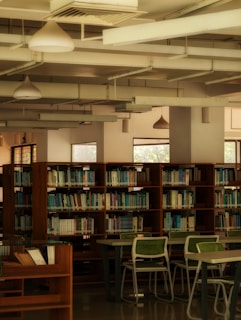 A library interior with wooden bookshelves filled with various books. The room features several tables and chairs with green cushions. Natural light enters through large windows, creating a warm and quiet atmosphere. The ceiling displays modern lighting fixtures and exposed ductwork.