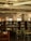 A library interior with wooden bookshelves filled with various books. The room features several tables and chairs with green cushions. Natural light enters through large windows, creating a warm and quiet atmosphere. The ceiling displays modern lighting fixtures and exposed ductwork.
