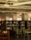 A library interior with wooden bookshelves filled with various books. The room features several tables and chairs with green cushions. Natural light enters through large windows, creating a warm and quiet atmosphere. The ceiling displays modern lighting fixtures and exposed ductwork.