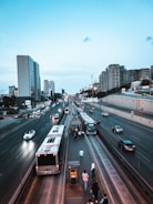 A bustling urban scene featuring a multi-lane highway with several buses and cars in motion. People are walking along an elevated pedestrian path situated between the lanes. High-rise buildings line the road on both sides under a clear blue sky.