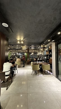 A modern restaurant interior with several people dining at tables. The ceiling is dark textured, and the lighting gives a warm and cozy ambiance. The seating consists of upholstered chairs and wooden tables. The decor includes wooden elements and indoor plants. There are large windows along one side, allowing natural light to enter during the day.