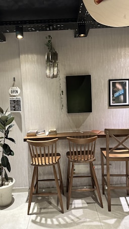 A cozy indoor space with a wooden counter and three wooden stools. There is a potted plant on the left and a small open sign with business hours next to it. A shelf or narrow table is adorned with a few books. On the wall, a piece of art displays a coffee-related theme, and a small decorative plant hangs above. The area is well-lit with overhead spotlights.