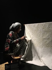 A person with gray hair and glasses is carefully working on a large piece of white fabric with intricate designs. The person is crouched and appears focused on the detailed artwork. The fabric is draped over a wooden frame, and there is a small bowl in front of the person, possibly containing tools or materials for the craft.