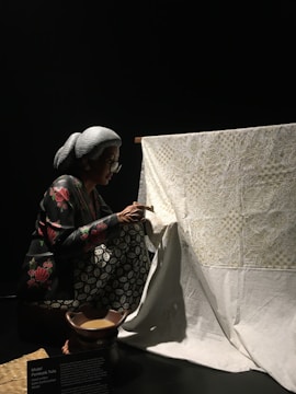 A person with gray hair and glasses is carefully working on a large piece of white fabric with intricate designs. The person is crouched and appears focused on the detailed artwork. The fabric is draped over a wooden frame, and there is a small bowl in front of the person, possibly containing tools or materials for the craft.