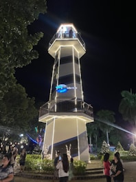 A tall, illuminated lighthouse with white stripes stands prominently at night. People are gathered around, some taking pictures. The surrounding area includes trees and shrubs adorned with lights, contributing to a festive atmosphere. The lighthouse sign reads 'The Lakeshore.'