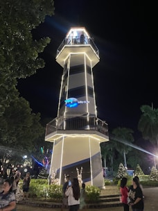 A tall, illuminated lighthouse with white stripes stands prominently at night. People are gathered around, some taking pictures. The surrounding area includes trees and shrubs adorned with lights, contributing to a festive atmosphere. The lighthouse sign reads 'The Lakeshore.'