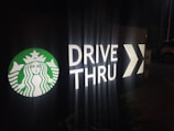 A dimly lit drive-thru sign features the Starbucks logo in green and white next to the words 'DRIVE THRU' in large illuminated letters, followed by a directional arrow.