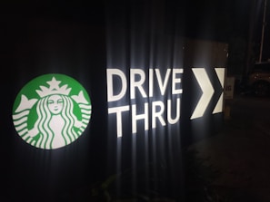 A dimly lit drive-thru sign features the Starbucks logo in green and white next to the words 'DRIVE THRU' in large illuminated letters, followed by a directional arrow.