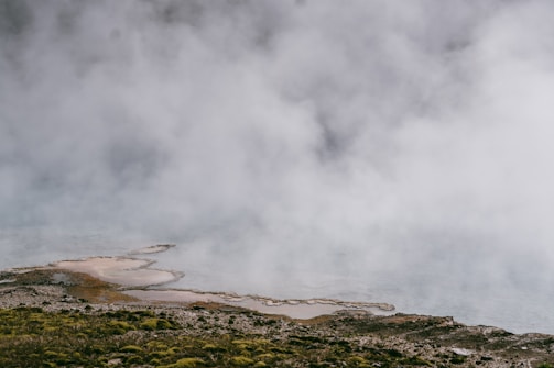 Dense mist or fog hovers over a textured landscape featuring patches of water and mossy terrain. The ground appears uneven, transitioning from rocky to vegetative surfaces as it extends into a body of water. The overall atmosphere is cool and mysterious.