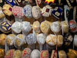 A collection of intricately designed traditional hats, adorned with ornate embroidery and vibrant patterns. The hats feature various embellishments such as beads, sequins, and tassels in a variety of colors including red, blue, gold, and white. They are displayed in a well-organized manner, showcasing their detailed craftsmanship.