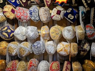 A collection of intricately designed traditional hats, adorned with ornate embroidery and vibrant patterns. The hats feature various embellishments such as beads, sequins, and tassels in a variety of colors including red, blue, gold, and white. They are displayed in a well-organized manner, showcasing their detailed craftsmanship.