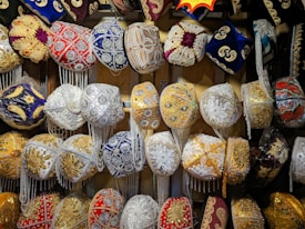 A collection of intricately designed traditional hats, adorned with ornate embroidery and vibrant patterns. The hats feature various embellishments such as beads, sequins, and tassels in a variety of colors including red, blue, gold, and white. They are displayed in a well-organized manner, showcasing their detailed craftsmanship.