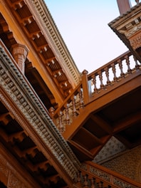 An image showcasing intricate wooden architecture, including detailed carvings and multiple levels of patterned balustrades. The structure is composed of rich brown wood with an open sky visible in the background, highlighting the craftsmanship and elegance of the design.