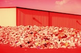 A large pile of assorted trash, including plastic bottles, bags, and various discarded items, is seen in front of an industrial building. The entire scene is bathed in a red hue, likely due to the lighting or color editing.