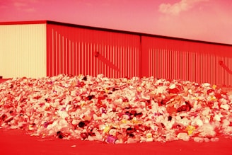 A large pile of assorted trash, including plastic bottles, bags, and various discarded items, is seen in front of an industrial building. The entire scene is bathed in a red hue, likely due to the lighting or color editing.